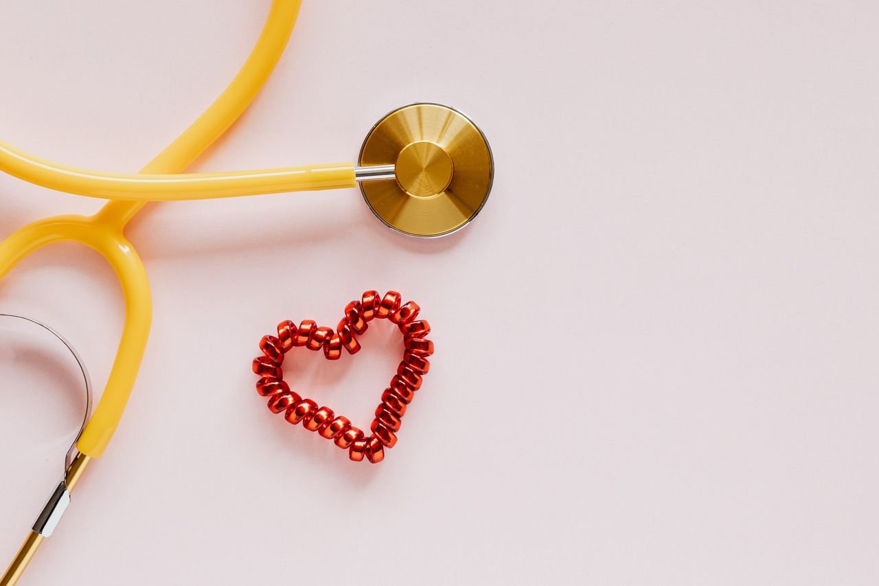 From above of yellow stethoscope near small red bead heart made for San Valentines Day on pale pink surface